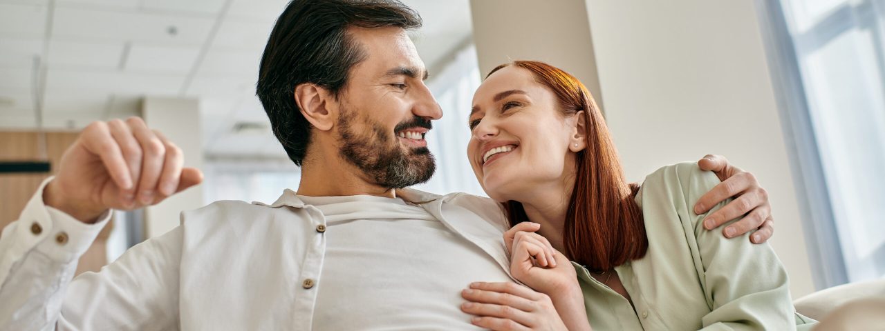 A beautiful adult couple, a redhead woman and a bearded man, sit together on a modern sofa in their apartment, enjoying quality time.